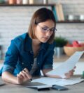 young woman working with computer while consulting some invoices and documents in the kitchen