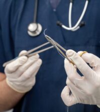 Doctor holds, in hand with disposable glove, forceps and needle holder and suture thread.