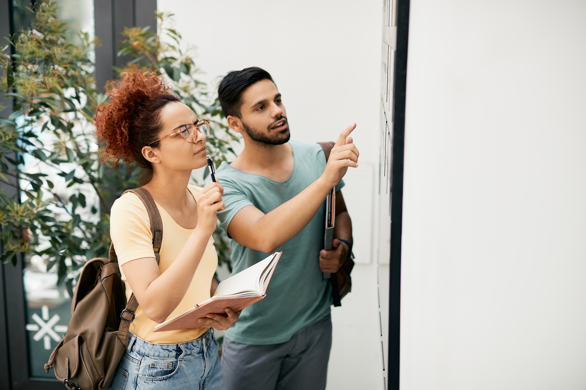 Two college students reading schedule of lectures on noticeboard in hallway.